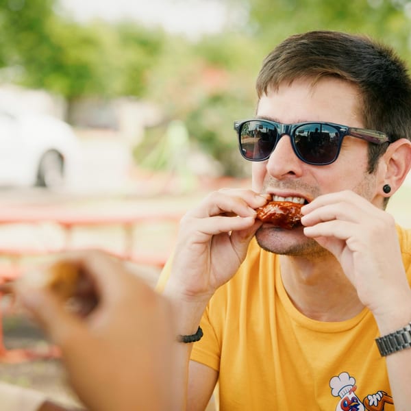 Man in sunglasses and yellow shirt eating a chicken wing outdoors at a picnic table.
