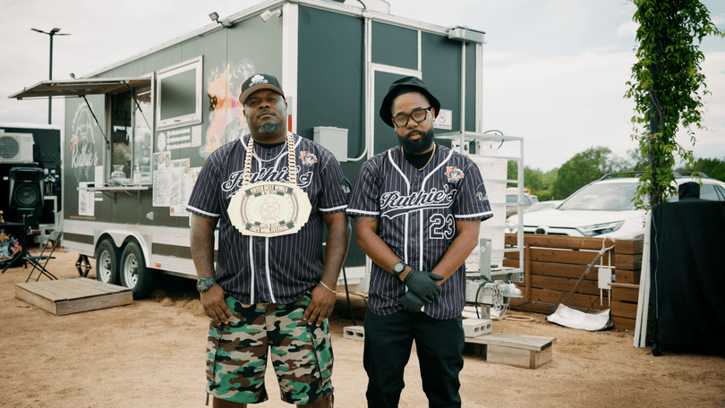 Two men in Ruthie's baseball jerseys stand in front of a Ruthie's food truck.