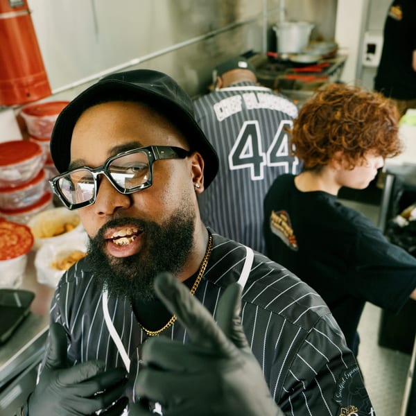 A man in a food truck kitchen, wearing glasses and a hat, gives a thumbs up.