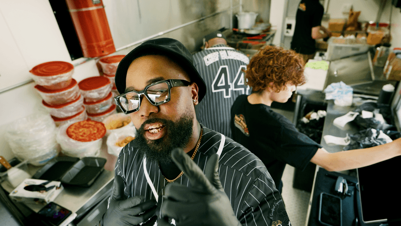 A man in a food truck kitchen, wearing glasses and a hat, gives a thumbs up.