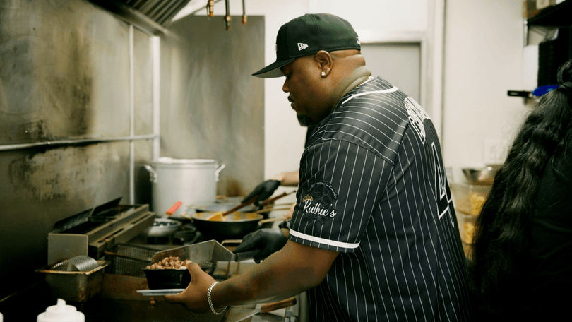 A chef in a Ruthie's branded shirt prepares food in a busy commercial kitchen.