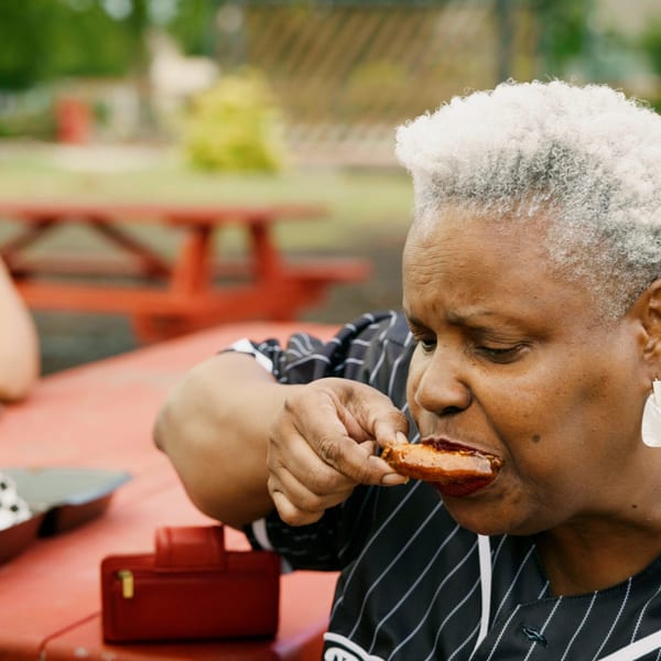 Woman with gray hair eating a rib outdoors at a red picnic table.