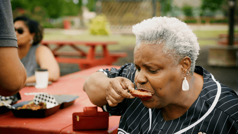 Woman with gray hair eating a rib outdoors at a red picnic table.