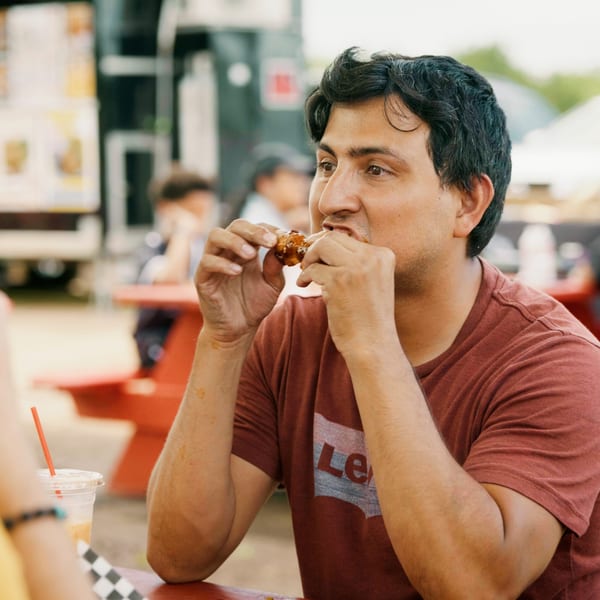 Man in a red shirt eating a chicken wing at an outdoor food truck park.