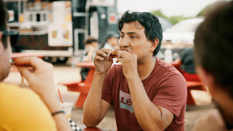 Man in a red shirt eating a chicken wing at an outdoor food truck park.