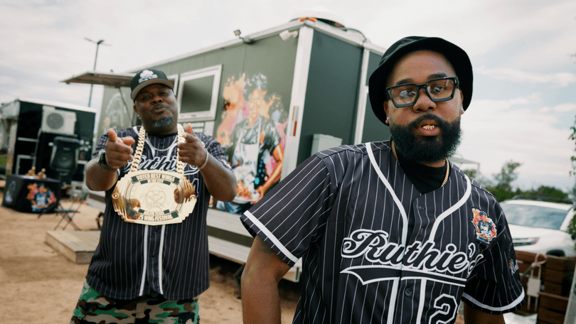 Two men in Ruthie's baseball jerseys pose in front of a food truck, one wearing a large gold chain.