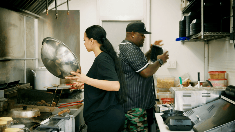 Two chefs, a woman and a man, working in a busy commercial kitchen with stainless steel equipment.