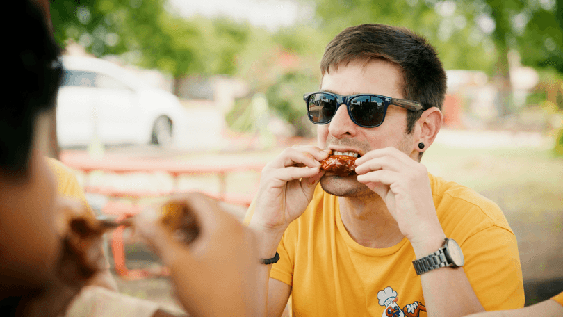 Man in sunglasses and yellow shirt eating a chicken wing outdoors at a picnic table.